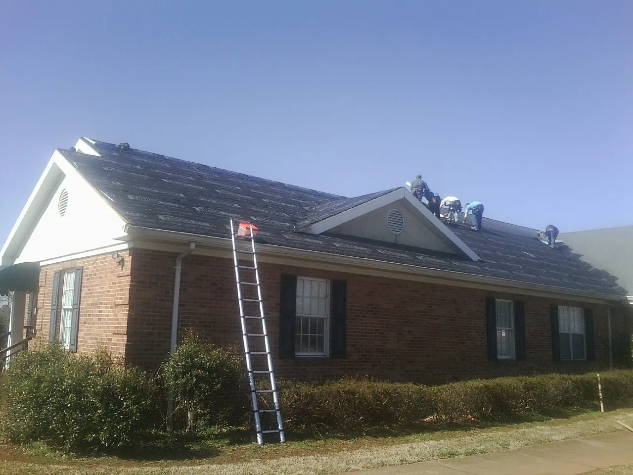 Crew tearing shingles off a brick ranch roof with a ladder set — Emergency Roof Repair work by our crew in Laingsburg
