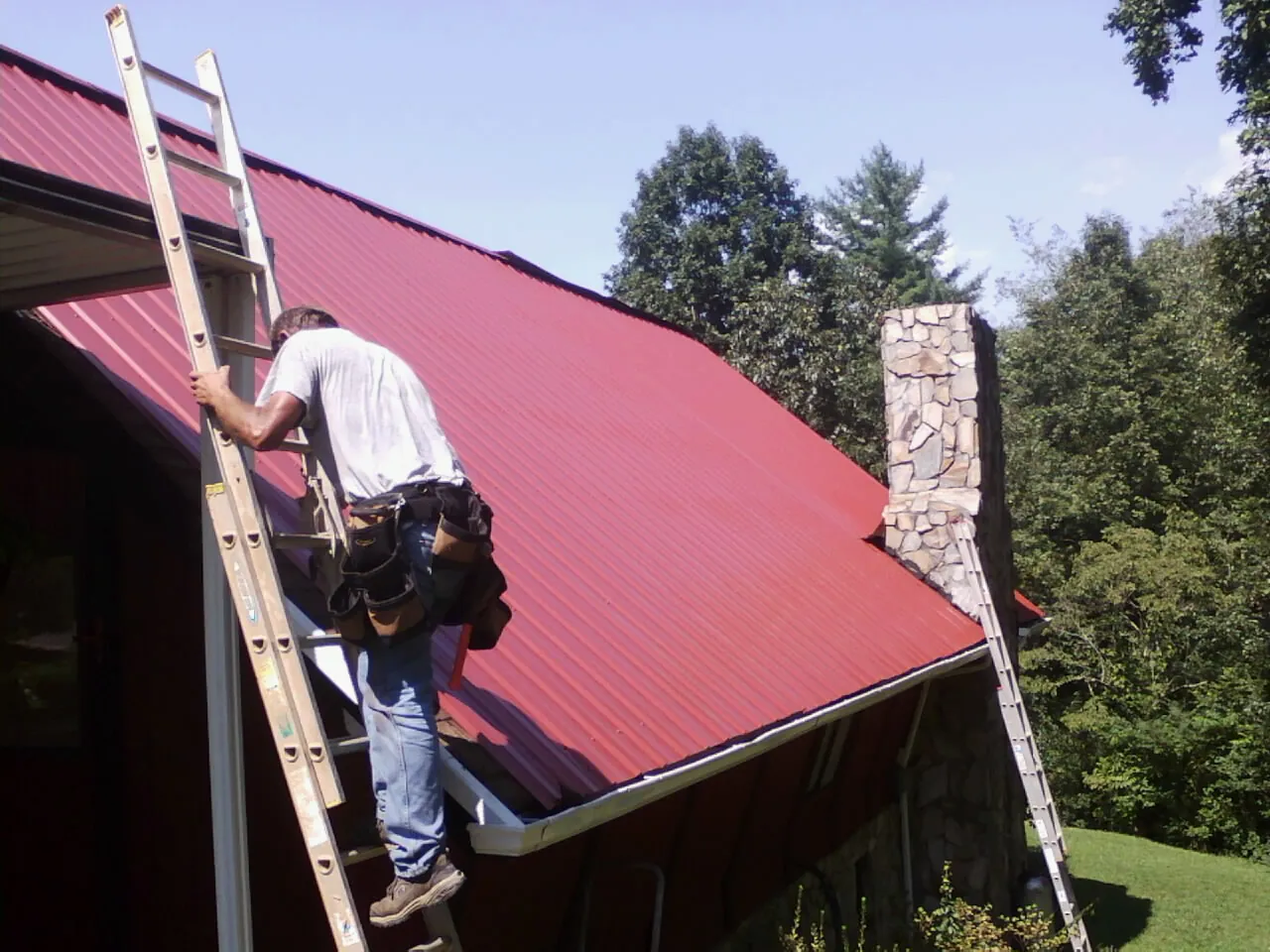 Roofer on a ladder beside a red metal roof and stone chimney — Soffit & Fascia Repair by Roofing Brothers in Laingsburg